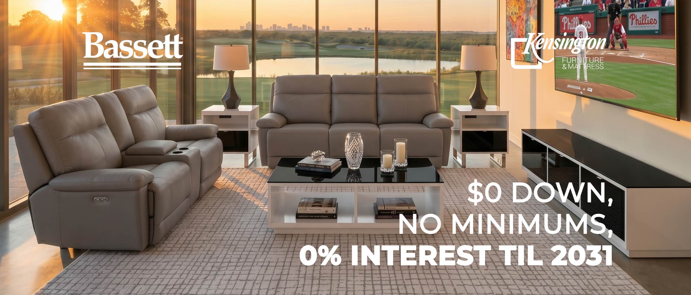 A modern living room scene showing a gray reclining loveseat and a matching three-seat sofa in front of large floor-to-ceiling windows at sunset. The room includes side tables, lamps, a glass coffee table, and a TV mounted on the wall.